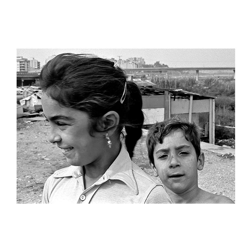Somriures entre runes  Una noia i un nen al campament del Riu Sec, entre barraques i ponts d’autopista. La noia somriu, com si per un instant oblidés el context que l’envolta. Any: 1982 – Fotografia de Vicenç Semper