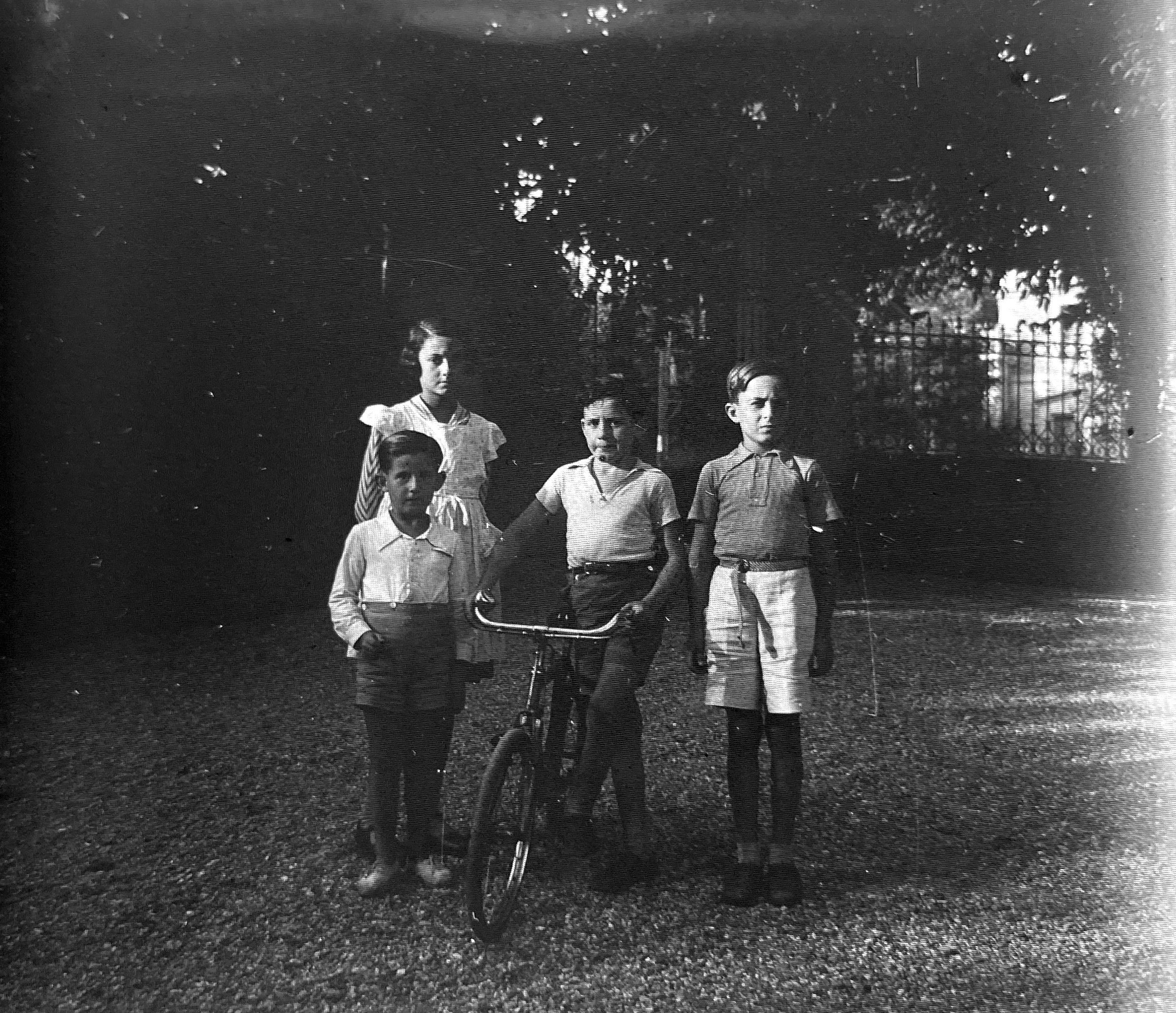 Quatre infants posen davant d’una bicicleta, en un entorn ombrívol i tancat amb reixa, símbol de les amplies propietats on estiuejaven famílies benestants com els Recolons. FOTO: Família Recolons / @GREC