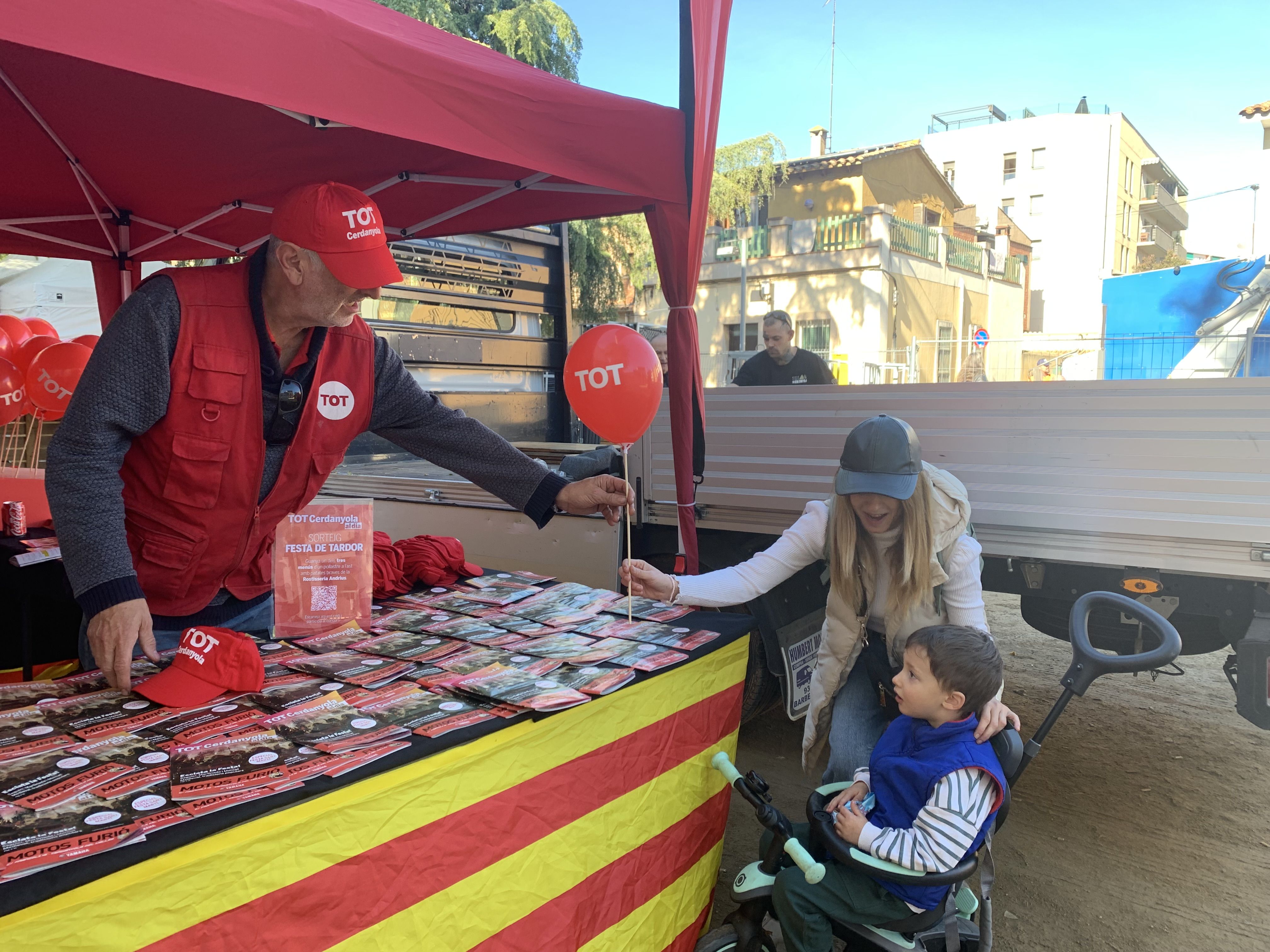 Veïns i veïnes s’acosten a la parada del TOT Cerdanyola durant la Festa de Tardor per endur-se revistes, globus i gorres.