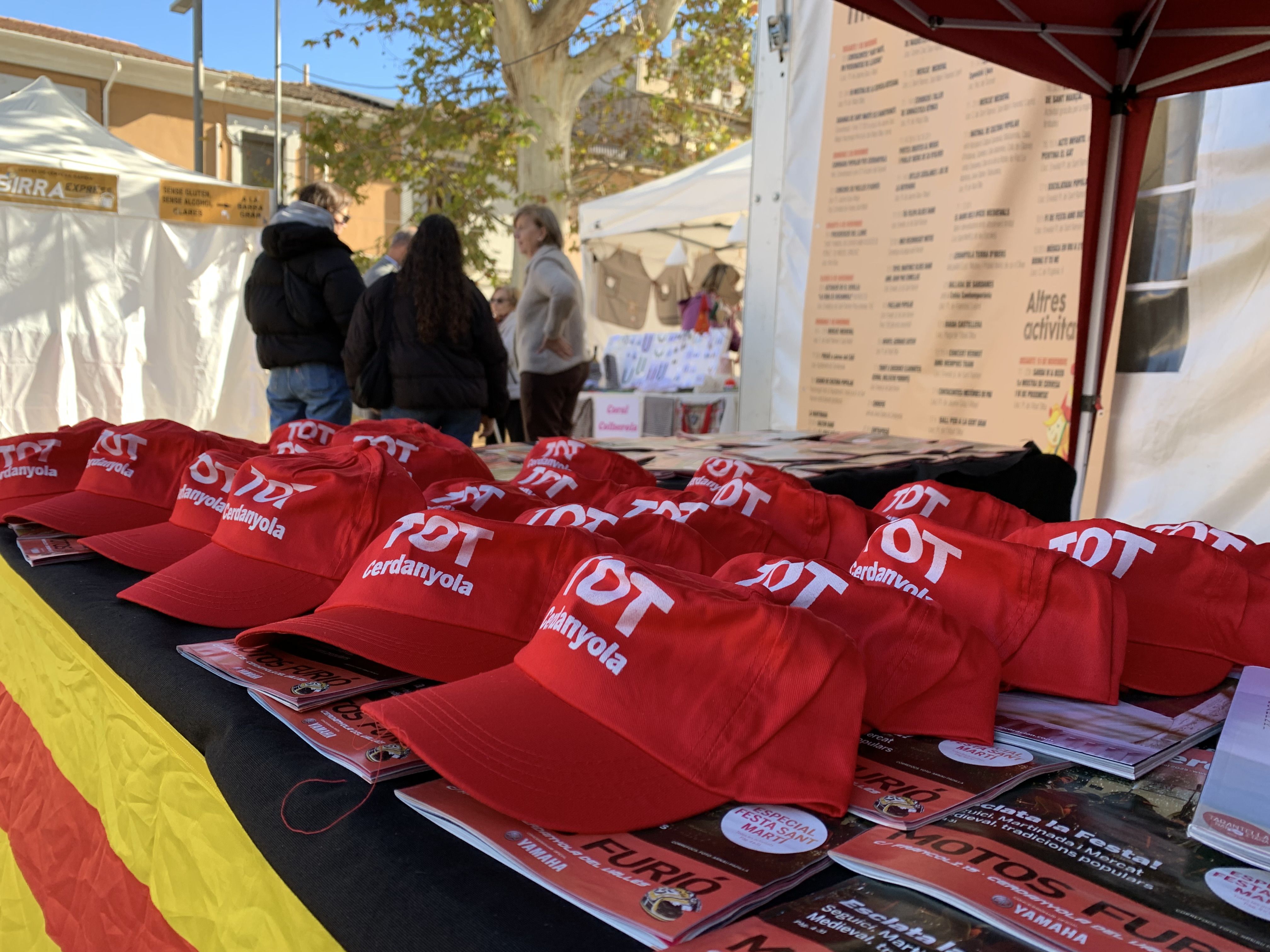 Veïns i veïnes s’acosten a la parada del TOT Cerdanyola durant la Festa de Tardor per endur-se revistes, globus i gorres.