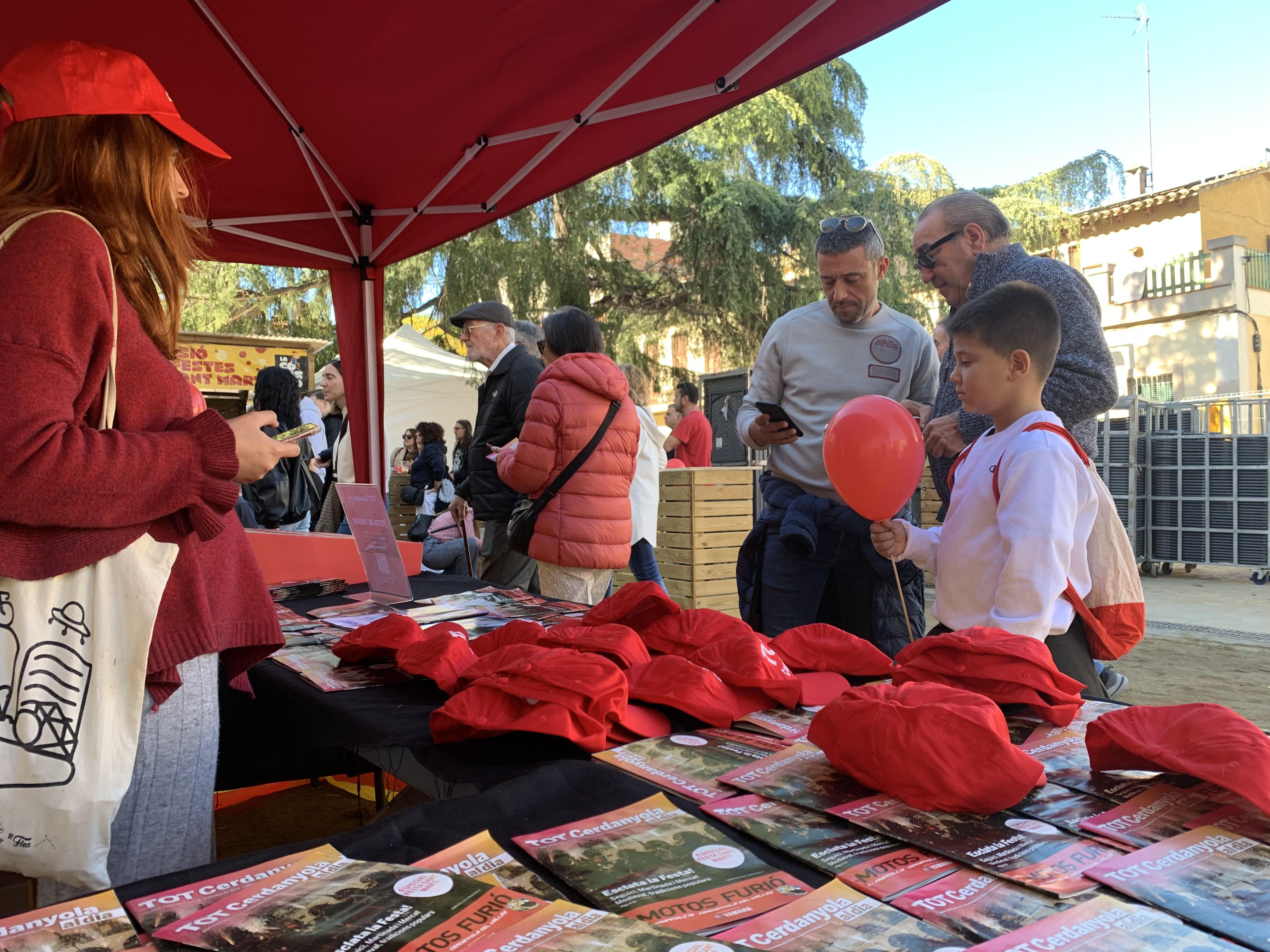 Veïns i veïnes s’acosten a la parada del TOT Cerdanyola durant la Festa de Tardor per endur-se revistes, globus i gorres.