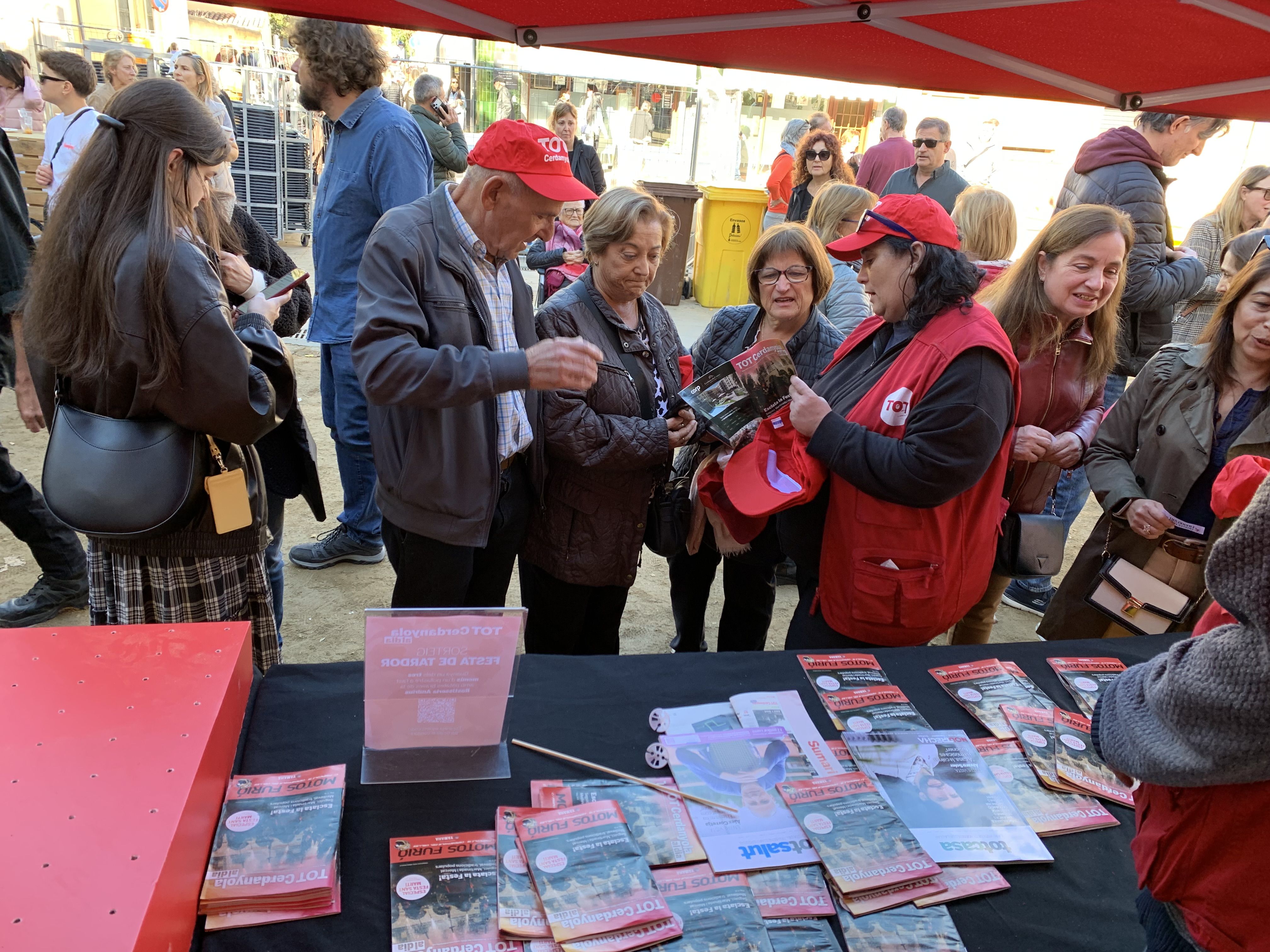 Veïns i veïnes s’acosten a la parada del TOT Cerdanyola durant la Festa de Tardor per endur-se revistes, globus i gorres.