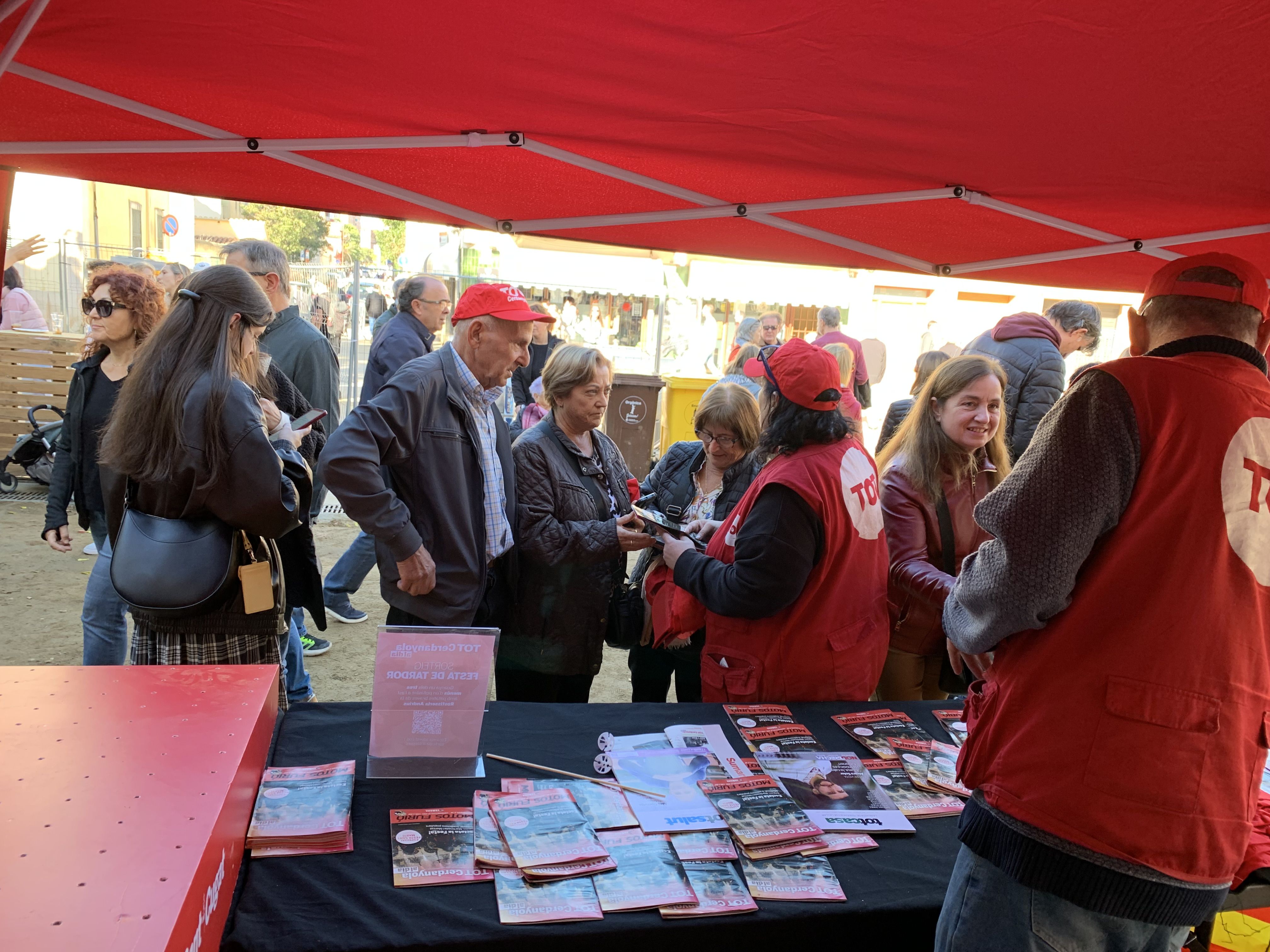 Veïns i veïnes s’acosten a la parada del TOT Cerdanyola durant la Festa de Tardor per endur-se revistes, globus i gorres.