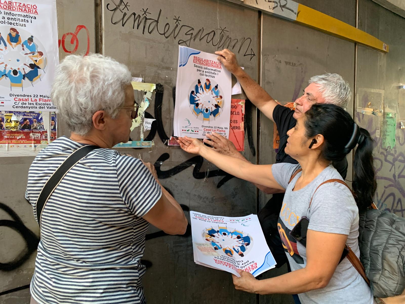 Voluntaris d’Acollim Cerdanyola-Ripollet enganxen cartells de la sessió informativa sobre la regularització extraordinària que es farà aquest divendres a La Flama. FOTO: Judit Josa